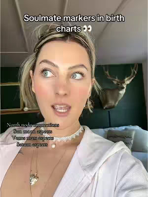 A woman is looking to the left and slightly upwards, with her mouth slightly open. She has light skin, blonde hair, and is wearing a white top with delicate necklaces. Behind her is a blurred background with what appears to be a deer head mounted on the wall.