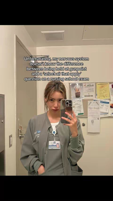 A woman in a grey nursing uniform takes a mirror selfie with her phone. Her brown hair is pulled back. The text overlay describes the stress of nursing school exams. Behind her is a beige wall with informational posters. The overall setting appears to be a nursing station or similar.