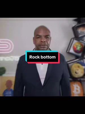 Image features a bald Black man in a dark blazer over a white shirt, standing in front of a neutral backdrop. A text overlay reading 'Rock bottom' in a white font on a black and red background is centered at the bottom of his chest. Behind him, there are various objects, including what looks like decorations of crypto logos and a neon sign.