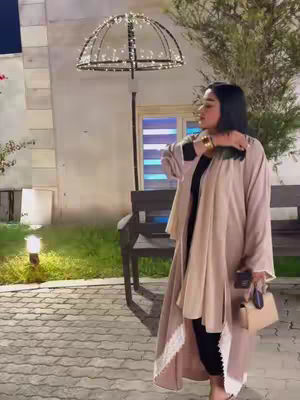 A woman stands outdoors at night, dressed in a light brown overcoat over a black outfit. She holds a small purse and a phone. The background includes a lit archway and a bricked pathway.