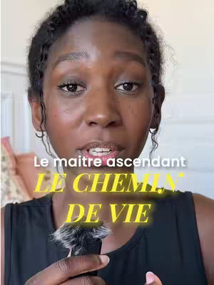 A woman with dark skin and curly hair is centered in the frame. The background is a lightly colored wall, with a pink item on the left side. The main subject is holding a microphone with a fuzzy black cover, with text overlayed on the image.