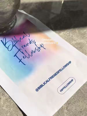 The image is an overhead shot of a page with watercolor strokes in shades of blue, purple, and orange dominating the upper portion. Overlaid in a handwritten-style dark blue font are the words 'Biblical friends Fellowship'. Below this the text '@BIBLICALFRIENDFELLOWSHIP' and '#BFFELLOWSHIP' are visible. A portion of a glass object is visible in the top left corner.