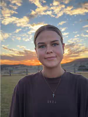 A young woman stands in front of a sunset backdrop, with a field and fence in the background. The woman is wearing a brown t-shirt and a silver necklace, with a cross pendant. The sky is filled with orange and blue hues.