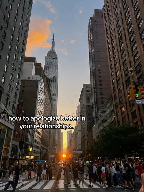 A photograph captures a bustling street scene in New York City, with the Empire State Building prominently featured at the end of the avenue. Buildings line both sides of the street, and a large crowd of people are seen walking on a crosswalk. The sky is a blend of orange and blue hues, suggesting a sunset. The overall composition is dynamic, with the buildings converging towards the Empire State Building, which serves as a focal point.