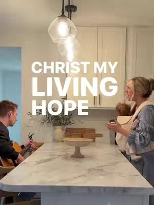 A well-lit kitchen scene with three people visible. The foreground features a large, light-colored countertop with a pedestal cake stand in the center. A man playing a guitar on the left, a woman holding a baby in a carrier on the right, and text overlayed in the center.