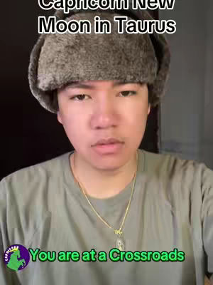 A person wearing a winter hat and a plain shirt is looking directly at the camera. The top text says 'Capricorn New Moon in Taurus' and at the bottom are the words 'You are at a Crossroads'. There is a logo on the bottom left.