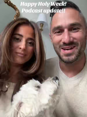 Two people are in a selfie-style shot with a dog in the foreground. The woman on the left has long brown hair and is smiling. The man is on the right and is also smiling and has short hair. A small, fluffy white dog is in front of them. The background includes a light fixture and a wall.