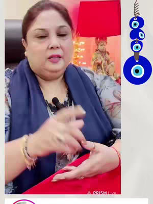 A woman is seated in front of what appears to be a video recording setup. She is wearing a blue shawl over a patterned top and a red dress. In the background, there's a lamp with a red lampshade and a small statue. A blue and white beaded ornament hangs to the right. She is gesturing with her hands as if speaking.
