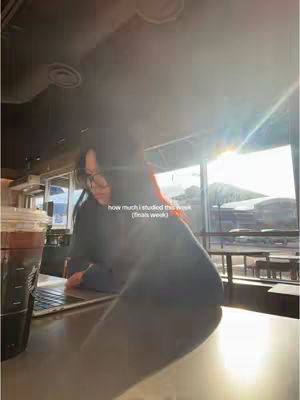 The image captures a study scene in a cafe environment. A person, likely a student, is seated at a table with a laptop and a large iced beverage, possibly coffee. The person is wearing glasses, and the scene is bathed in natural light from a window, suggesting a bright and airy atmosphere. Text overlays are present, indicating the video's theme possibly centered around the study habits or experiences.