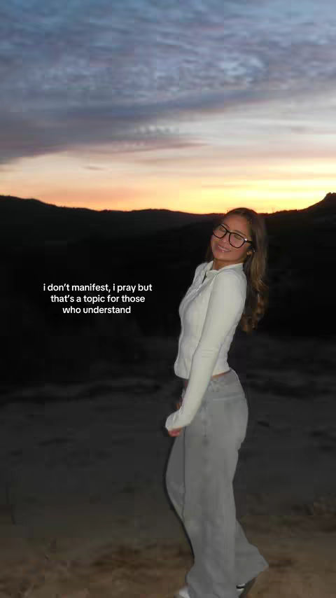 A young woman wearing glasses and a white jacket poses with a sunset in the background. Text overlayed on the image reads, "i don't manifest, i pray but that's a topic for those who understand".