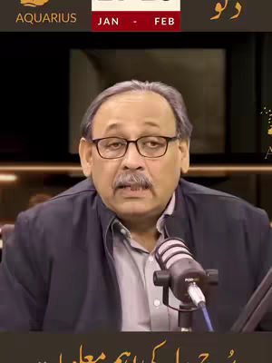 A man wearing glasses and a suit jacket is centered in the frame. A microphone is positioned in front of him.