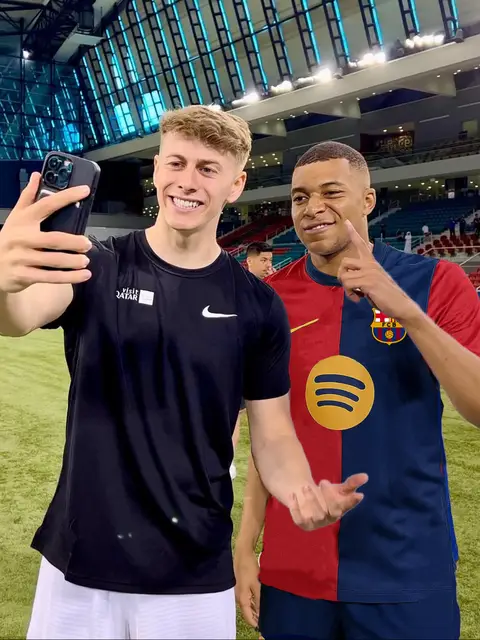The cover image is a selfie taken by a young man with short blonde hair, smiling and holding up a black smartphone to capture a shot with a dark-skinned man. The person on the right is pointing his finger towards his temple. They appear to be on a soccer field within an indoor sports facility. The background has seating and large, geometric ceiling panels. The man on the right is wearing a red, blue, and gold soccer jersey, while the selfie-taker is in a black t-shirt and white shorts.