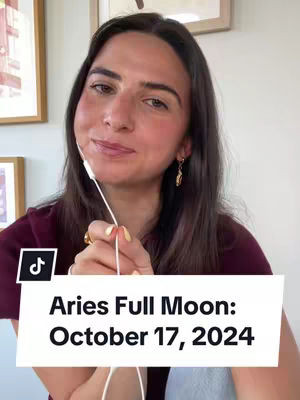 A woman with long dark hair is centered against a neutral background, likely indoors. She wears a maroon top and gold earrings. A white overlay at the bottom shows the date and event of the Aries Full Moon. The Tiktok logo is visible, along with the woman's face.