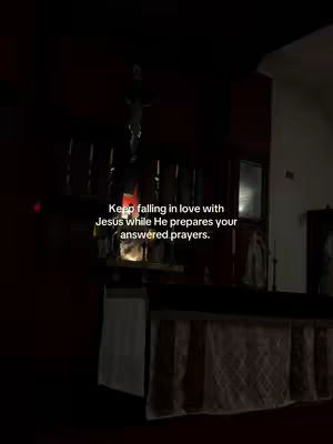 The image shows a dimly lit church interior with a crucifix hanging above a simple altar. Text overlayed with the sentence: Keep falling in love with Jesus while He prepares your answered prayers.