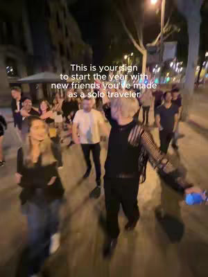A night scene captures a group of people walking on a cobblestone street, illuminated by streetlights. Text overlays suggest a message about starting the year with new friends made as a solo traveler. The people appear to be dressed casually, and the overall vibe is communal and welcoming.