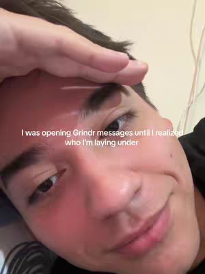A close-up shot of a young man's face, angled from a low perspective. The man has dark hair and fair skin, with a slight flush to his cheeks and nose. He's looking towards the lower right of the frame with a soft, almost smiling expression. A hand is positioned over his forehead. The background is a light, neutral color. Text is overlaid on the image.