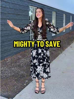 A woman with long dark hair and a black and white floral dress is standing outdoors with her arms outstretched to the sides. She is smiling. The background includes a building with dark gray siding and several windows, along with a patch of greenery and a paved pathway.