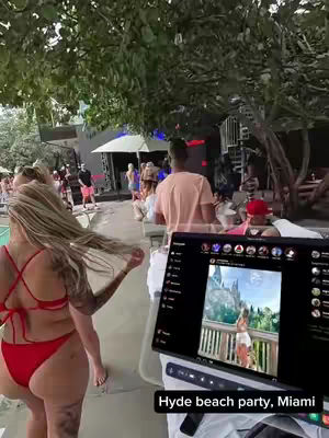 A woman in a red bikini stands with her back to the camera as she walks away from the viewer, while a man holds a laptop that displays a woman on a beach, with a party going on in the background. The image is taken outside with natural light.