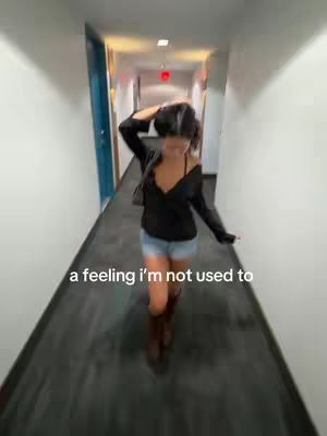 A woman walks down a hallway with white walls and a dark floor. She is wearing a black top and denim shorts. Text overlayed on the image reads "a feeling I'm not used to".