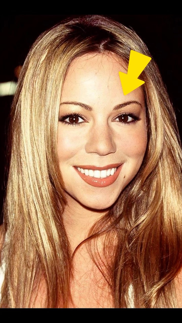 A close-up shot of a smiling woman, Mariah Carey, with long blonde hair. She has fair skin, brown eyes, visible mascara, and her lips are colored with a soft, peachy lipstick. The lighting is even, highlighting her facial features. A yellow arrow points towards her forehead.