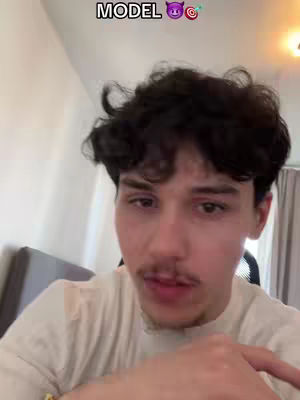 A selfie-style shot features a young man, centered in the frame, with dark, curly hair and a mustache. The text overlay "MODEL" is at the top left along with some emojis including two devil horns, and a target with an arrow through it. The man is looking directly at the camera with a slight smile, wearing a light-colored shirt. The background is slightly blurred, with a hint of an interior setting.