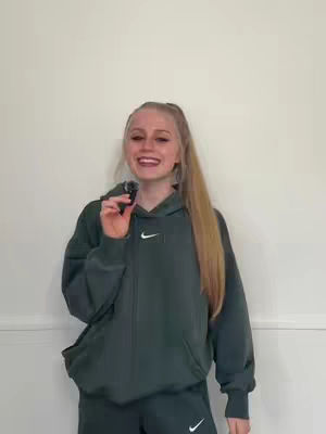 A young woman with blonde hair in a high ponytail holds a microphone and smiles at the camera while wearing a green Nike hoodie. She is set against a plain white background.