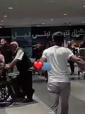 A scene at what appears to be an airport or train station, two men are in the foreground, one pushing a wheelchair, a figure, presumably the camera operator, is walking in an opposite direction. There are other people in the background with a sign in the backdrop.