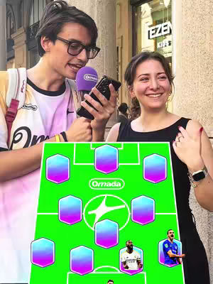 A man and a woman smiling, holding a purple microphone and a phone, respectively. In the foreground, a green football formation graphic is displayed.