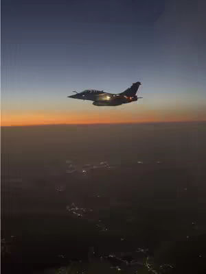 A high-angle shot of a jet fighter flying in the sky at sunset. The plane is silhouetted against a gradient of colors in the sky, from deep blue at the top to orange and yellow near the horizon. Below the horizon is a dark, undefined landmass with some visible lights, suggesting a city or populated area. The composition is vertical, with the aircraft occupying the central space and creating a sense of motion.