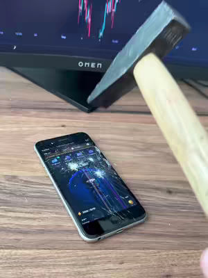 A phone with a damaged screen lies on a wooden surface. A hammer is positioned above it, suggesting an impending impact. A computer monitor with a financial chart is visible in the background.