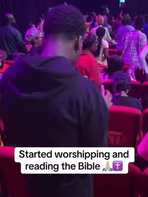 A person wearing a dark hoodie is seen from behind, facing forward. The background is filled with people seated in rows, with red seats and a purple-toned lighting. White text overlay at the bottom indicates it is a worship service.