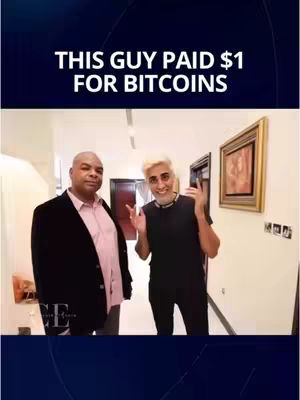 Two men stand in a well-lit hallway. The man on the left wears a black blazer over a pink shirt. The man on the right has light hair and is wearing a black shirt.