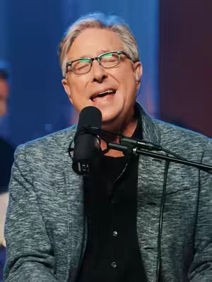 The cover image features an older man with glasses singing into a microphone. He is wearing a black shirt under a gray patterned blazer. The background is a blurred blue, and other microphone stands are visible. The man's expression is one of open-mouthed singing or speaking, with his eyes closed. The lighting appears to be soft, highlighting his face.