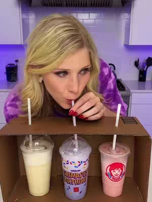 A woman with blonde hair is leaning over a cardboard box with three drinks in it. She has a straw in her mouth. The drinks in the box are a light yellow, a blue and white cup, and a pink drink. The background is of a kitchen.