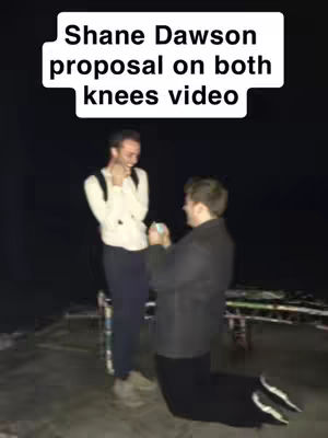 Two men are shown in an outdoor nighttime setting. One man is kneeling on one knee, holding a ring box, and looking up at the other man, who is smiling and appears surprised. The kneeling man is wearing a black jacket and pants, while the other man is wearing a white shirt and dark pants. Above them is a white text box that reads "Shane Dawson proposal on both knees video".