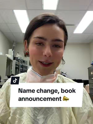 A woman with dark hair and fair skin looks at the camera and smiles. She appears to be in a medical or laboratory setting. The background is a bit blurry, showing equipment and lights on the ceiling. There is a white banner with the text "Name change, book announcement" and a speaker symbol.