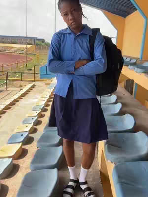 A young person stands at a stadium, wearing a blue school uniform and a backpack. They are positioned in front of rows of seats, with a slight smile on their face.