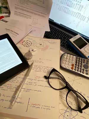 An overhead shot of a desk covered in study materials. There is a laptop open, a tablet, papers with handwritten notes and diagrams, pens, a calculator, and a pair of glasses. The objects are arranged in a neat, organized manner, suggesting a study setup.
