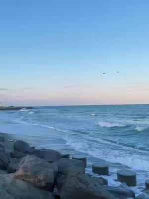 A serene seascape features a clear blue sky transitioning to a soft pink near the horizon. The ocean waves gently roll towards a rocky shore in the foreground. The overall composition evokes tranquility.