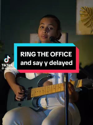 A person is playing a guitar while seated. The cover image features a person of color in a white top seated on a chair playing an electric guitar. Behind them, a blurred background gives depth. Overlaid text in a rectangular box reads 'RING THE OFFICE and say y delayed' in white text with a red and blue background, adding a communicative layer.