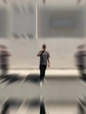 A man wearing a gray t-shirt, black pants, and a cap is walking towards the camera, presumably holding a phone to his ear in an outdoor setting. The blurred background suggests movement or a shallow depth of field. The composition has radial blur creating a motion effect.