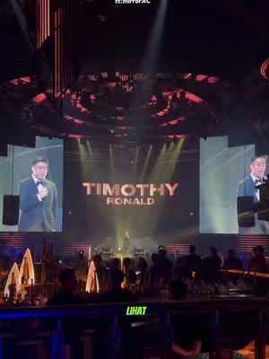 The cover photo captures a live presentation on a large stage with a man speaking. The background features the name of the presenter 'TIMOTHY RONALD' in prominent, golden letters on a large screen. Below, a large crowd is gathered, with spotlights illuminating the stage and background.