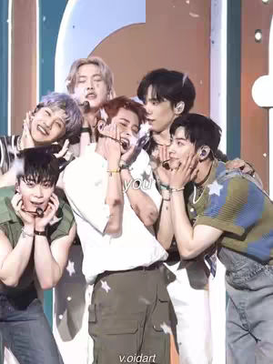 The cover image features seven young men in a close-up shot. They appear to be members of a K-pop group, posing and interacting with each other. The background is softly lit, suggesting a performance setting, with warm tones prevailing. Some members hold their hands over their faces or heads, conveying a sense of surprise or affection. The composition is framed tightly, focusing on the faces and upper bodies of the group members. The text overlay at the bottom right states the social media handle.