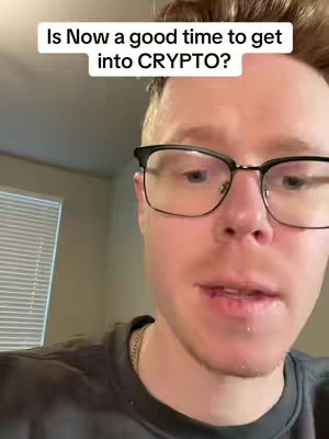 A man with short, light brown hair and glasses is centered in the frame. The background includes an indiscernible wall and a partly visible window with horizontal blinds. The man is wearing a black shirt. A text overlay asks, "Is Now a good time to get into CRYPTO?"