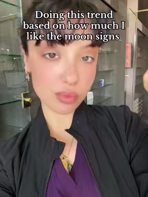 A selfie-style video cover image featuring a woman with dark hair and a nose ring. The woman is facing the camera, with text overlaying the image. The background suggests an indoor setting, possibly a shop or office, with shelves and items visible.