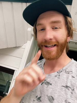 A man with ginger hair and a beard is pointing at the camera, wearing a cap and a patterned t-shirt, with a bright, engaging expression, set against a light-colored wall background.