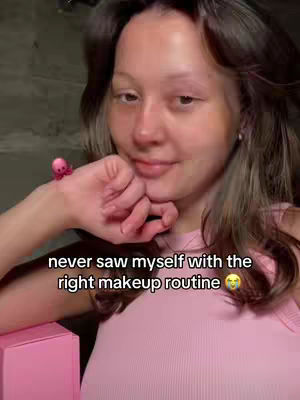 A woman with light skin and dark hair is positioned in a softly lit space. She is wearing a light pink tank top. Her arm is bent, and her hand gently touches her face near her chin. The woman's expression is serene and confident. A small, pink decorative piece resembling an octopus is attached to her wrist. In the lower left corner, a pink box adds another color element to the scene. The background appears blurred and out of focus.