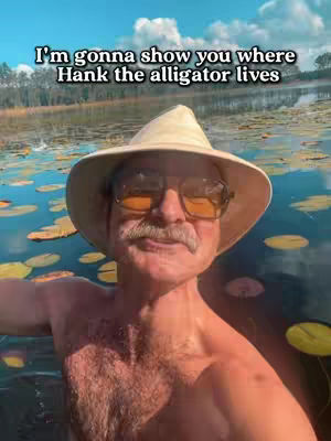 A man with a mustache and a hat stands in a pond. Text overlays say, 'I'm gonna show you where Hank the alligator lives.' The background shows a pond with lily pads and trees.