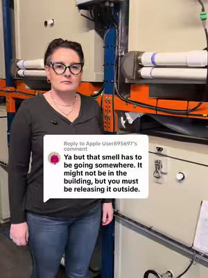 A woman wearing glasses and a black top stands indoors, in front of large metallic machinery with pipes and orange and blue structural elements visible. A white speech bubble overlays the image, containing overlaid text.