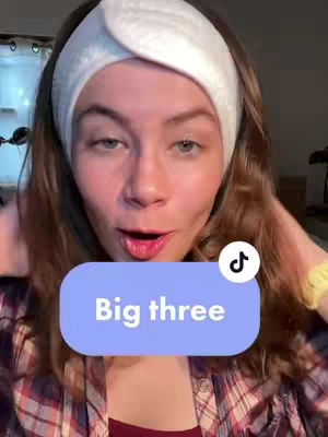A woman with fair skin and light brown hair looks directly at the camera, wearing a white spa headband and a plaid shirt. Overlayed on the image is a light blue rounded rectangle containing the text 'Big three'. In the upper right corner, a white and black TikTok logo. The background is out of focus, featuring curtains. The woman has her lips open, as if she is talking.